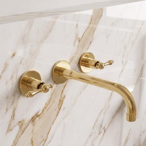 Puede incluir: Un grifo de baño montado en la pared con un acabado en oro pulido. El conjunto incluye un caño central curvado y dos manijas de palanca separadas, cada una montada sobre una placa base circular para controlar el flujo de agua.