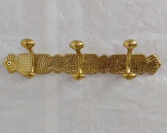 May include: A gold metal coat rack with three hooks. The rack has a textured surface and is mounted on a wall.