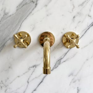 Peut inclure: Un ensemble de robinetterie de salle de bain en or poli comprenant un bec central courbé flanqué de deux poignées en croix. La robinetterie est fixée sur une surface en marbre blanc avec des veines grises, offrant un fini métallique contrasté.