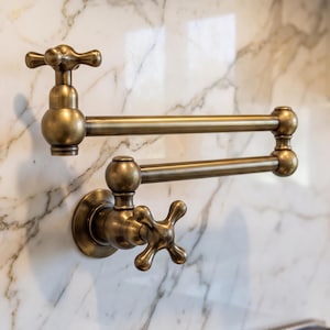 May include: A wall-mounted pot filler faucet with a brushed brass finish. This articulated fixture features two horizontal arms with swivel joints and two cross-style handles, one located at the base and one at the nozzle end for water control.