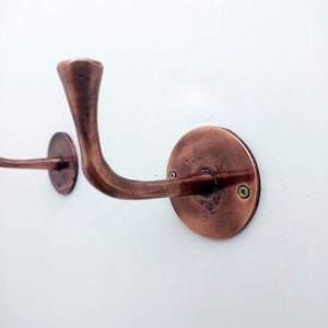 May include: Two copper wall hooks with a curved design and a round base. The hooks are mounted on a white wall.