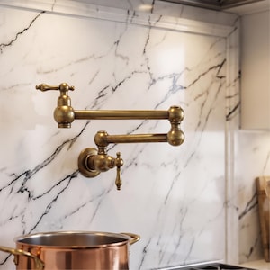 May include: A wall-mounted pot filler faucet with a brushed antique brass finish. The fixture features a double-jointed, articulating swing arm design with two separate lever handles for water control, mounted directly onto a white marble wall.