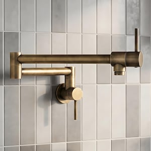 May include: A wall-mounted pot filler faucet with a brushed brass finish, featuring a double-jointed articulating swing arm, two cylindrical control handles, and a circular wall plate, designed for installation above a kitchen stove.