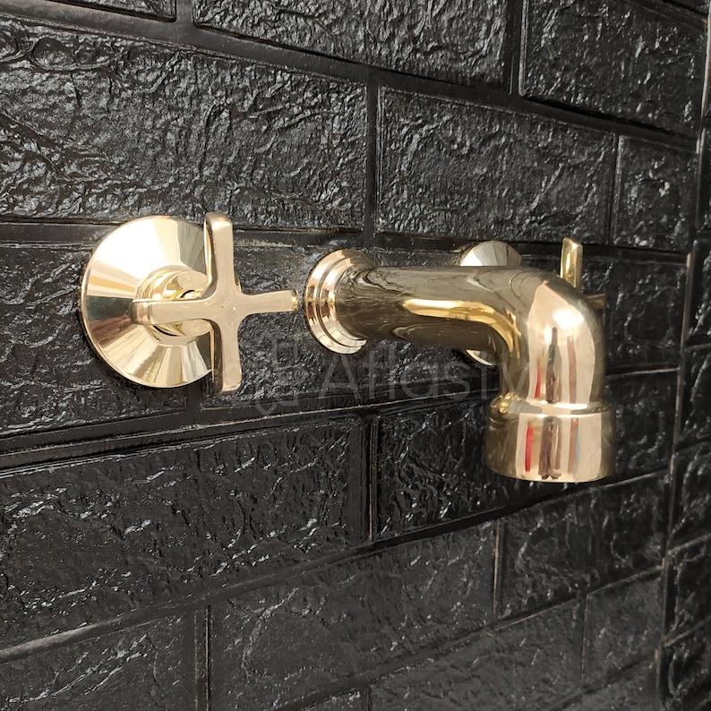 May include: A gold-toned faucet with a cross-shaped handle mounted on a black brick wall.