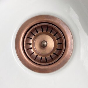 May include: A circular copper-toned sink drain strainer set into a white basin. The metal strainer features a central knob and a series of evenly spaced vertical slots arranged in a ring around the middle, designed for water drainage.