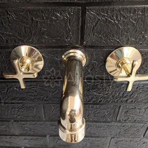 May include: Three gold-toned bathroom faucets mounted on a black tiled wall. The faucets have cross-shaped handles and are in a polished finish.