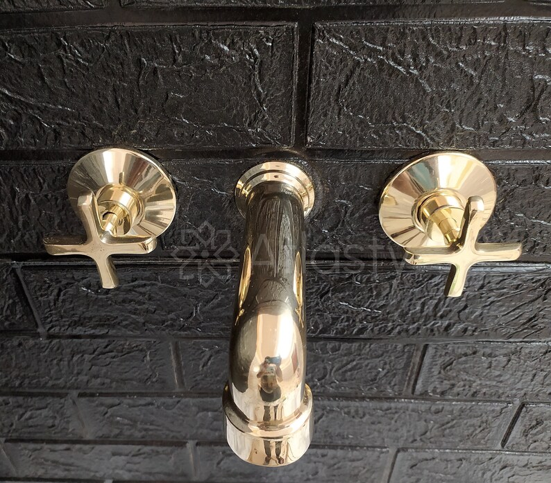 May include: Three gold-toned bathroom faucets mounted on a black tiled wall. The faucets have cross-shaped handles and are in a polished finish.
