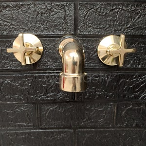 May include: A gold-toned bathroom faucet with two cross-shaped handles. The faucet is mounted on a black tiled wall.