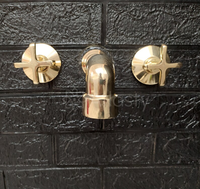 May include: A gold-toned bathroom faucet with two cross-shaped handles. The faucet is mounted on a black tiled wall.