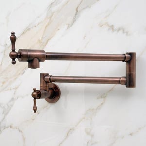 Puede incluir: Un grifo de pared para llenar ollas con un diseño de brazo oscilante articulado de doble articulación. El accesorio presenta un acabado de cobre cepillado oscuro y dos manijas de palanca verticales para el control del agua, extendiéndose horizontalmente.