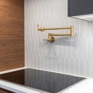 Op de afbeelding: Een aan de muur gemonteerde, scharnierende pannenvul-kraan met een geborstelde gouden afwerking, voorzien van twee horizontale zwenkarmen en een enkele bedieningshendel, geïnstalleerd boven een zwarte inductiekookplaat tegen witte tegels.