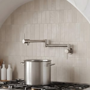 May include: A brushed nickel wall-mounted pot filler faucet with a dual-jointed swing arm and two vertical lever handles, positioned above a large stainless steel stockpot resting on a black gas stovetop with heavy-duty metal burner grates.