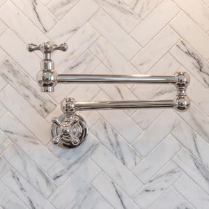 May include: A polished chrome wall-mounted pot filler faucet featuring a double-jointed articulating arm and two cross-style handles. The fixture has a reflective metallic finish and is mounted against a white marble tile wall with a herringbone pattern.