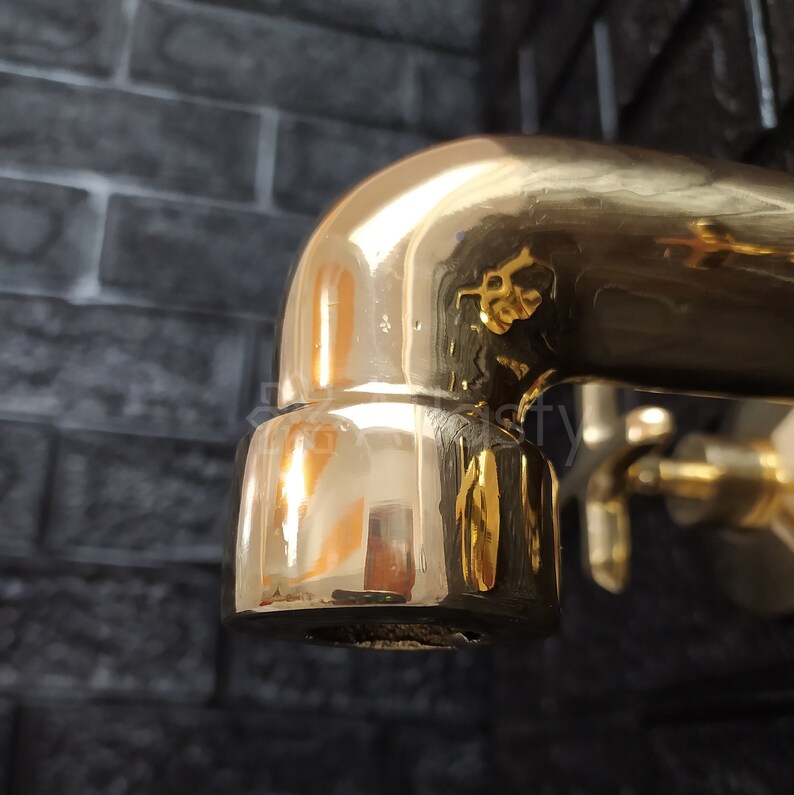 May include: Close-up of a gold-toned faucet spout with a shiny, reflective surface. The faucet is against a backdrop of dark gray brick.