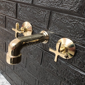May include: A gold-toned bathroom faucet with two cross-shaped handles mounted on a black brick wall.