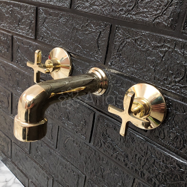May include: A gold-toned bathroom faucet with two cross-shaped handles mounted on a black brick wall.