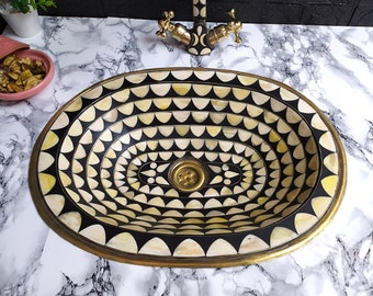Op de afbeelding: Ovaalvormige badkamer wastafel met een zwart-wit geometrisch patroon. De wastafel is gemaakt van messing en heeft een gouden afwerking. De wastafel is omgeven door een wit marmeren aanrecht.