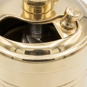 May include: A close-up of a brass pepper grinder. The grinder has a cylindrical body with a curved top and a small handle. The brass has a polished finish, reflecting light.