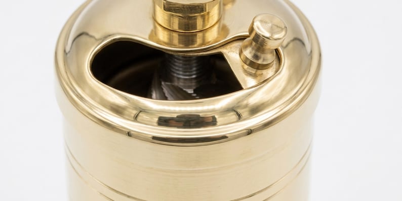 May include: A close-up of a brass pepper grinder. The grinder has a cylindrical body with a curved top and a small handle. The brass has a polished finish, reflecting light.