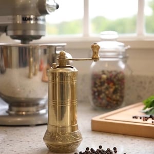 May include: A brass pepper mill with a hand crank sits on a countertop. The mill is cylindrical with decorative detailing. A stainless steel stand mixer and a glass jar filled with peppercorns are in the background. Fresh tomatoes and herbs are also visible.