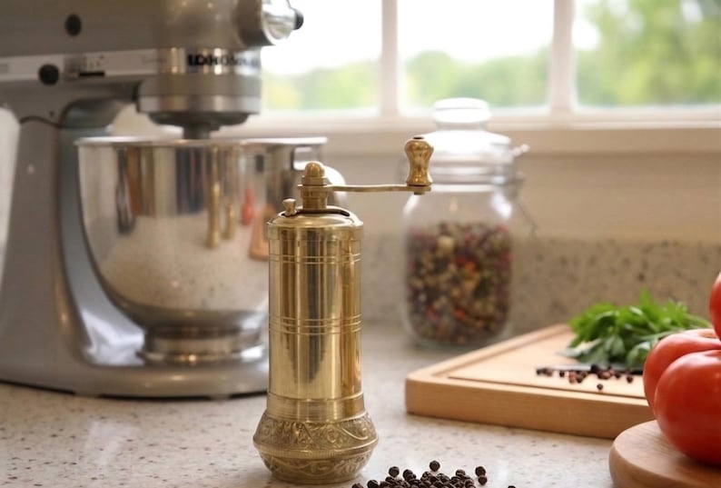 May include: A brass pepper mill with a hand crank sits on a countertop. The mill is cylindrical with decorative detailing. A stainless steel stand mixer and a glass jar filled with peppercorns are in the background. Fresh tomatoes and herbs are also visible.