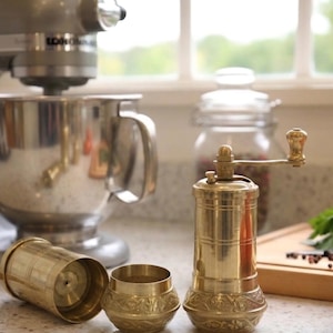 May include: A kitchen scene featuring a silver stand mixer, a brass pepper grinder, and a small brass container. A glass jar with peppercorns, tomatoes, and fresh herbs are also visible. The grinder is approximately 6 inches tall.