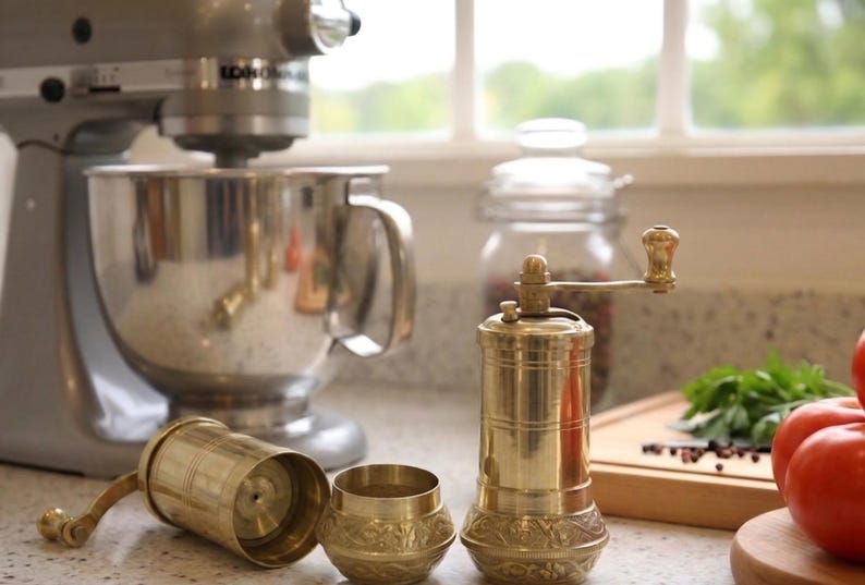 May include: A kitchen scene featuring a silver stand mixer, a brass pepper grinder, and a small brass container. A glass jar with peppercorns, tomatoes, and fresh herbs are also visible. The grinder is approximately 6 inches tall.