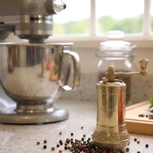 May include: A silver stand mixer with a stainless steel bowl is next to a brass pepper grinder. The grinder has a decorative base and a wooden handle. Scattered peppercorns and a jar of spices are also visible.