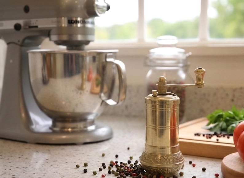 May include: A silver stand mixer with a stainless steel bowl is next to a brass pepper grinder. The grinder has a decorative base and a wooden handle. Scattered peppercorns and a jar of spices are also visible.