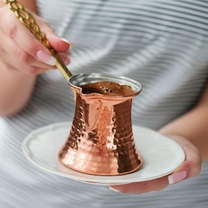 May include: A copper Turkish coffee pot with a hammered finish, filled with coffee, on a white saucer. The pot is being held by a hand with a gold spoon.