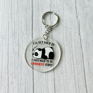 May include: A round, clear acrylic keychain with a black and white panda sleeping on it. The text "I'll get over it I just need to be dramatic first" is printed on the keychain.