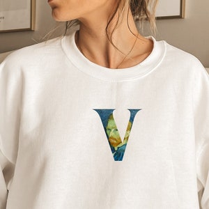 May include: White sweatshirt with a large, stylized letter "V" printed on the front. The letter is filled with a painting of a man's face, reminiscent of a famous artist's work.