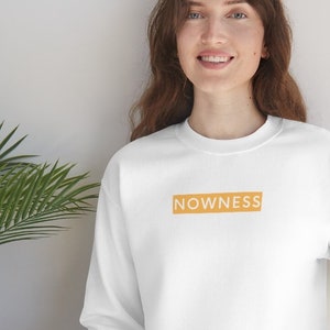 Op de afbeelding: Witte trui met een lichtoranje doosje met daarop "NOWNESS".