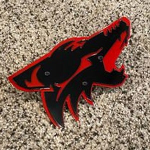 May include: A black and red wolf head emblem with a howling expression. The emblem has a glossy finish and is mounted on a textured, speckled surface. The wolf's silhouette is black, with a red outline and interior.