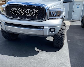 Puede incluir: Una camioneta plateada con una parrilla de malla negra que dice "CUMMINS" en letras blancas. La camioneta tiene neumáticos grandes y está estacionada frente a un garaje.