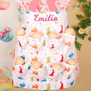 May include: A white apron with pockets filled with eggs, featuring a colorful bunny pattern. The apron has the name "Emilia" embroidered in pink. The top of the apron is attached to a pink floral top with a large pink bow.