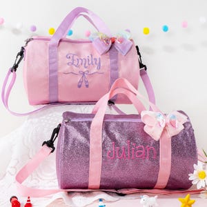 May include: Two glittery duffel bags, one pink and one purple, with matching straps and bows. The pink bag has "Emily" embroidered on it, and the purple bag has "Julian". Both bags have a ballet theme.