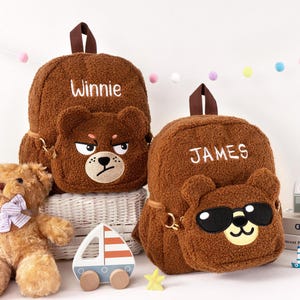 May include: Two brown teddy bear-themed backpacks with the names "Winnie" and "JAMES" embroidered on them. One bear has an angry face, the other wears sunglasses. A teddy bear, a wooden sailboat, and a star are in the foreground.