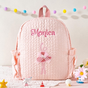 May include: A light pink backpack with a textured pattern, featuring the name "Monica" embroidered in pink script. The backpack has a ballerina design and side pockets. A pink handle is at the top.