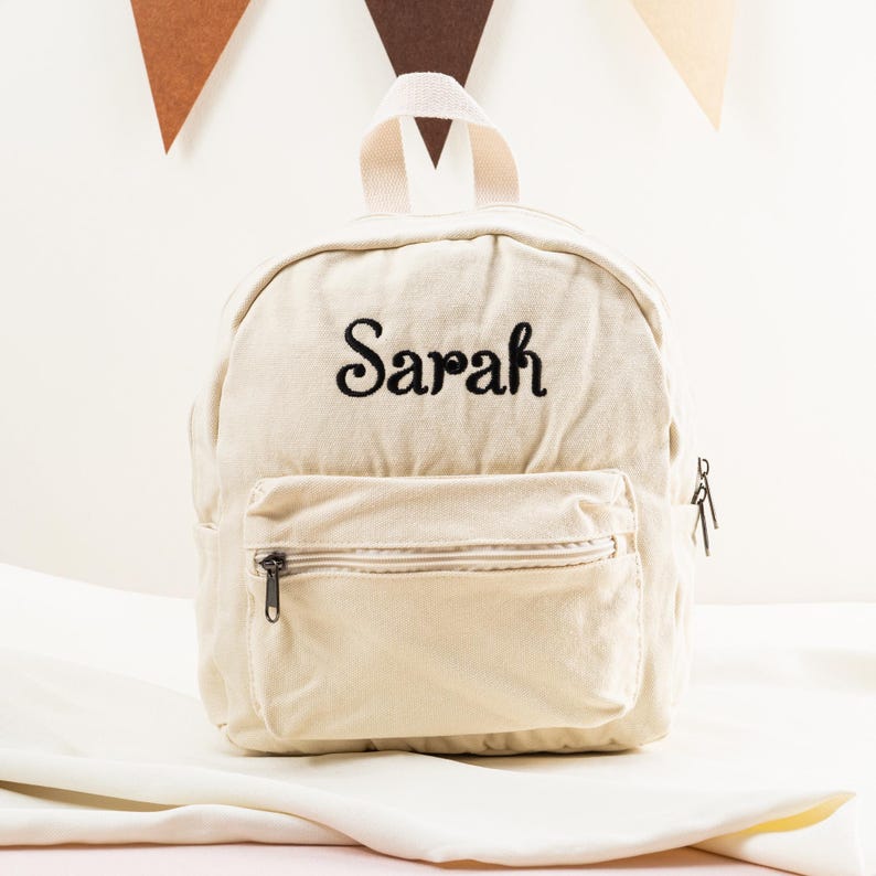 K&ouml;nnte beinhalten: Ein cremefarbener Canvas-Rucksack mit dem Namen "Sarah", der in Schwarz auf der Vorderseite gestickt ist. Der Rucksack hat eine Rei&szlig;verschlusstasche auf der Vorderseite und einen Rei&szlig;verschluss oben.