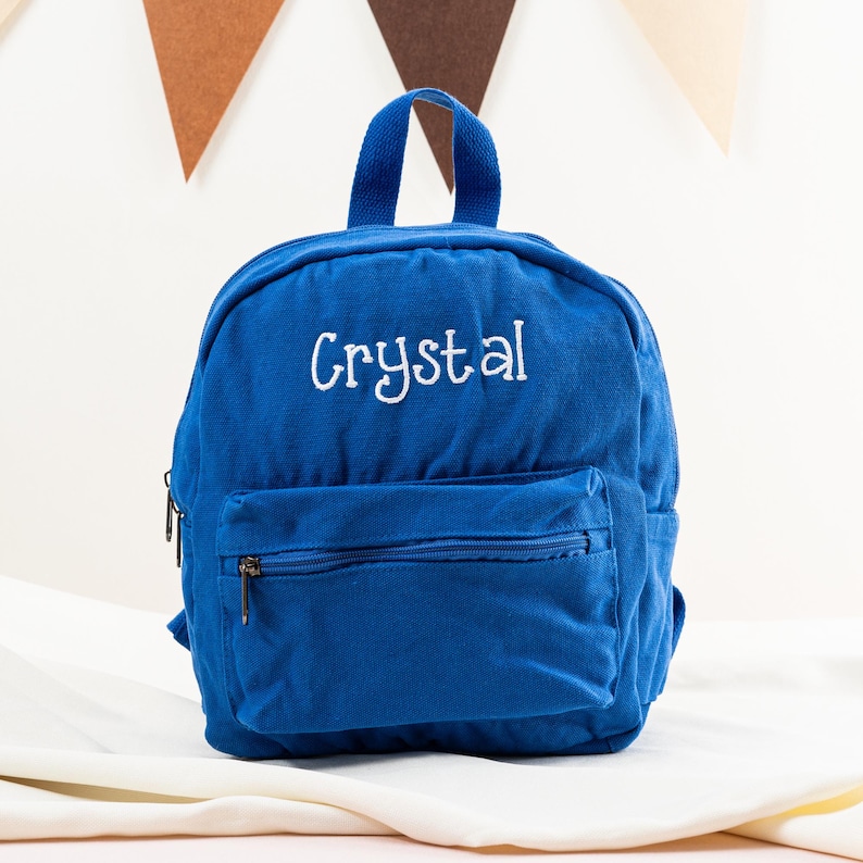 K&ouml;nnte beinhalten: Ein blauer Canvas-Rucksack mit dem wei&szlig;en gestickten Namen "Crystal" auf der Vorderseite. Der Rucksack hat eine Rei&szlig;verschlusstasche auf der Vorderseite und verstellbare Tr&auml;ger.