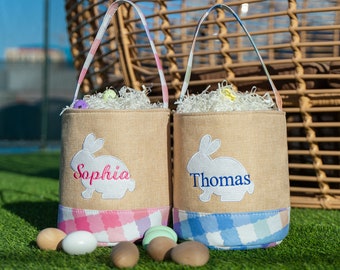 Può includere: Due cestini pasquali in juta con bordo a quadri rosa e blu. I cestini sono personalizzati con i nomi "Sophia" e "Thomas" e presentano una silhouette di coniglio bianco. I cestini sono riempiti con carta triturata e uova di Pasqua.