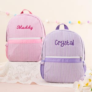 May include: Two personalized backpacks, one pink with white stripes and the name "Maddy" embroidered on it, and one purple with white stripes and the name "Crystal" embroidered on it.