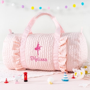 Puede incluir: Una bolsa de deporte rosa claro con una superficie texturizada y bordes con volantes. La bolsa presenta un bordado de bailarina y el nombre "Melissa" en escritura rosa. Tiene dos asas y una cremallera. Hay juguetes en miniatura en primer plano.