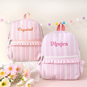 May include: Two pink and white gingham backpacks with embroidered names. One backpack has "Crystal" embroidered in orange, and the other has "Monica" embroidered in pink. Both have a ruffle detail and a top handle.