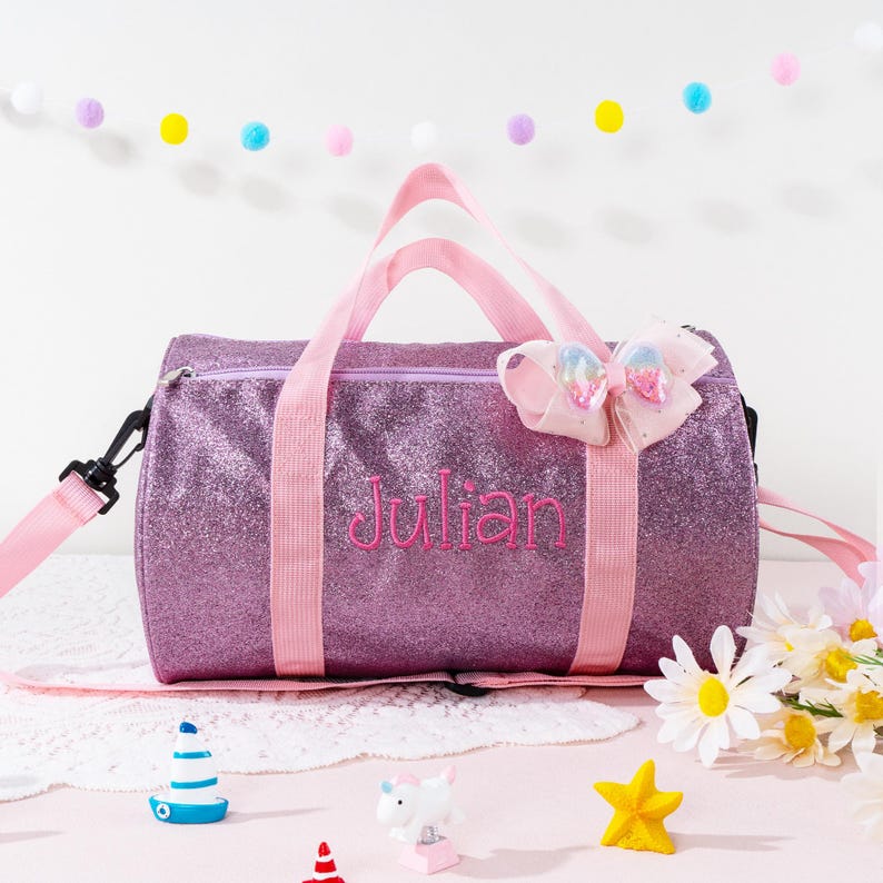 May include: A purple glittery duffel bag with pink nylon handles and an adjustable shoulder strap. The name Julian is embroidered in pink thread on the front. A decorative pink bow with iridescent accents is attached to the top right side.