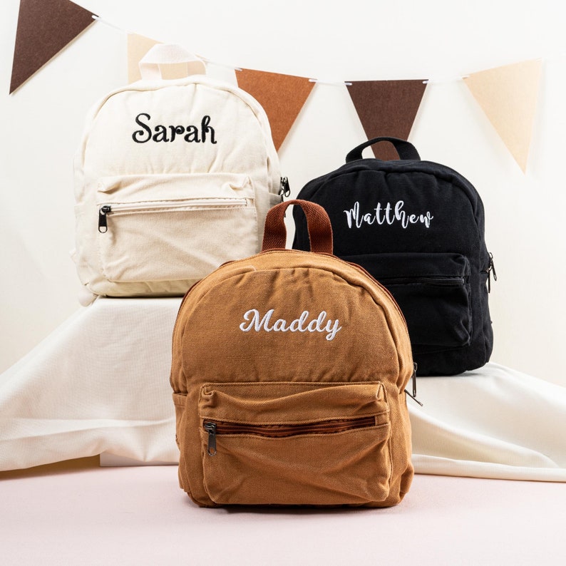 K&ouml;nnte beinhalten: Drei personalisierte Mini-Rucks&auml;cke in Beige, Braun und Schwarz mit den Namen "Sarah", "Maddy" und "Matthew", die in Wei&szlig; gestickt sind.