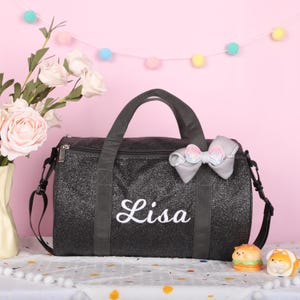May include: A black glittery duffel bag featuring the name Lisa embroidered in white script on the front. The bag includes two black fabric handles, a detachable shoulder strap, and a decorative grey bow with iridescent sequins attached to the side.
