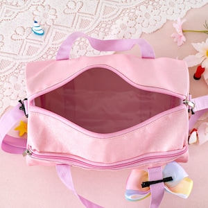 May include: A light pink duffel bag with a wide, open zippered main compartment and a smaller zippered front pocket. The bag features light purple fabric handles and an adjustable shoulder strap, accented with a multi-colored pastel ribbon bow.