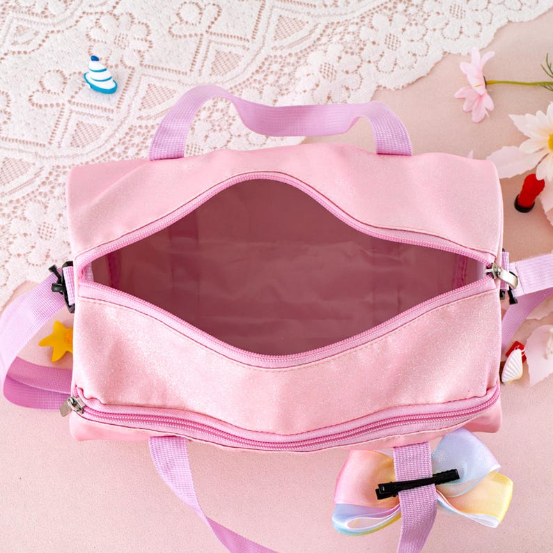 May include: A light pink duffel bag with a wide, open zippered main compartment and a smaller zippered front pocket. The bag features light purple fabric handles and an adjustable shoulder strap, accented with a multi-colored pastel ribbon bow.